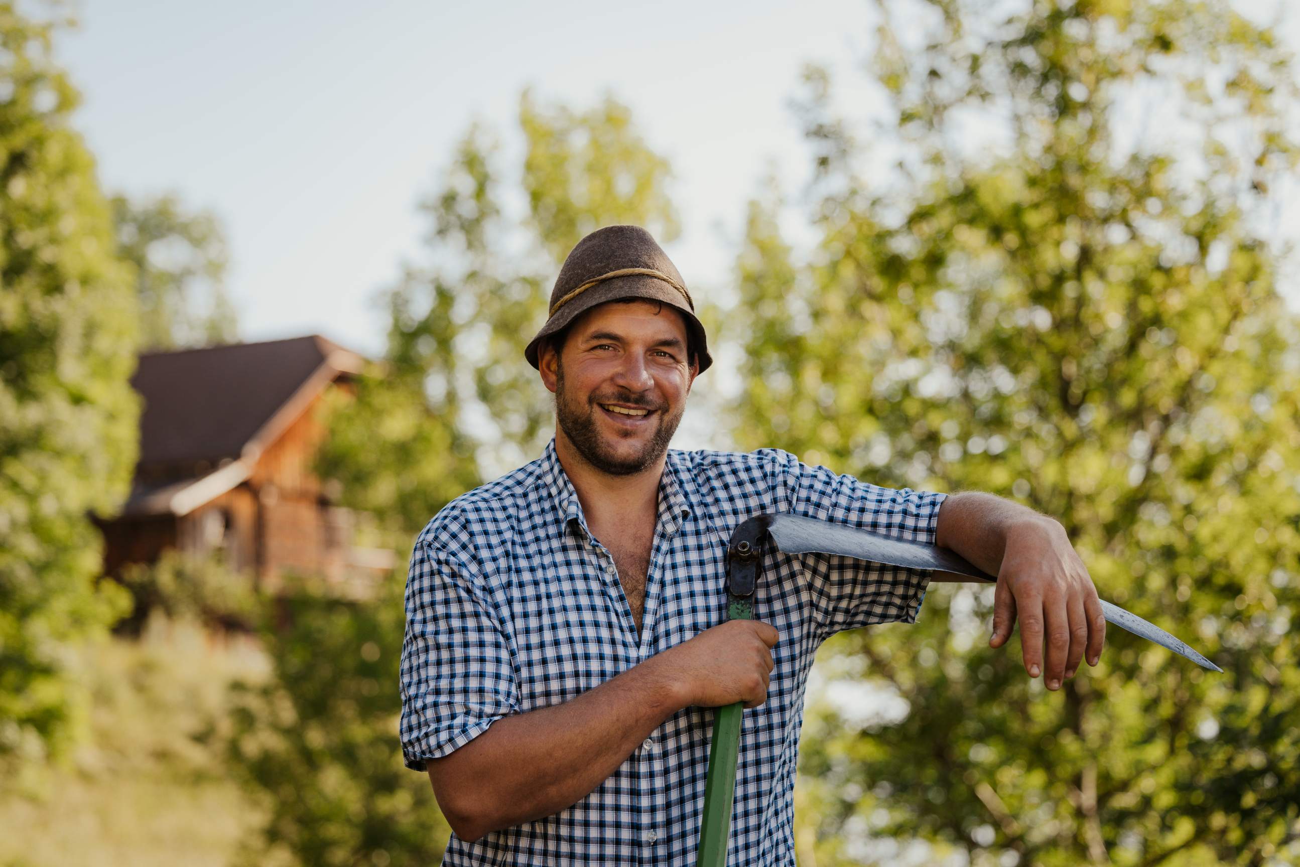 Ein lächelnder Bauer mit Hut und kariertem Hemd hält eine Sense in der Hand.