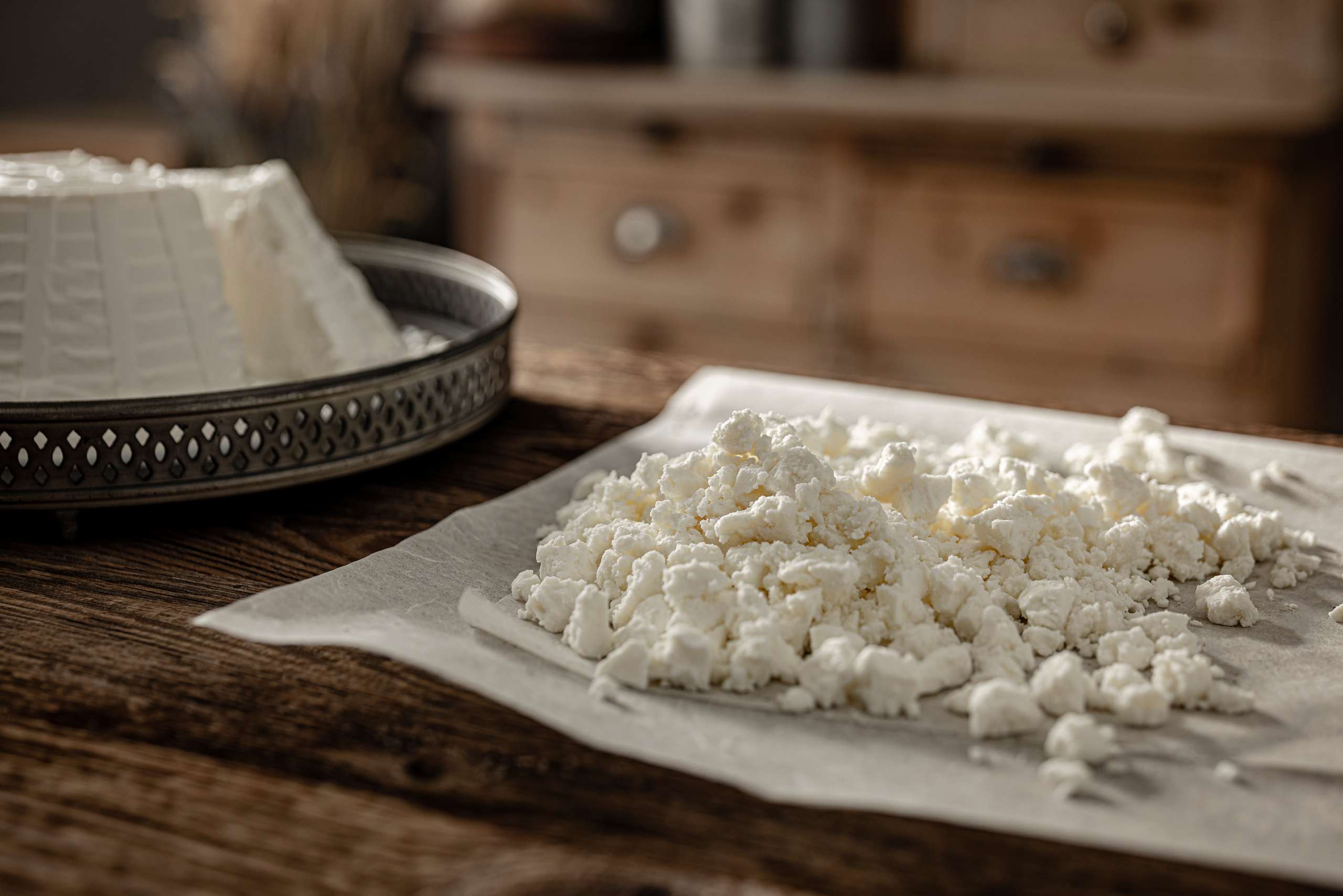 Frisch zerbröselte Ricotta auf Backpapier, mit einer ganzen Form im Hintergrund einer rustikalen Küche.
