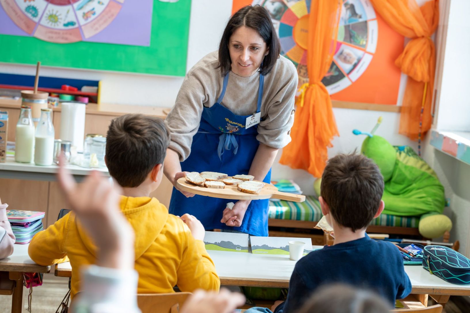Frau mit blauem Schurz reicht Kindern im Klassenzimmer Brote mit Milch-Aufstrich auf einem Holzbrett.