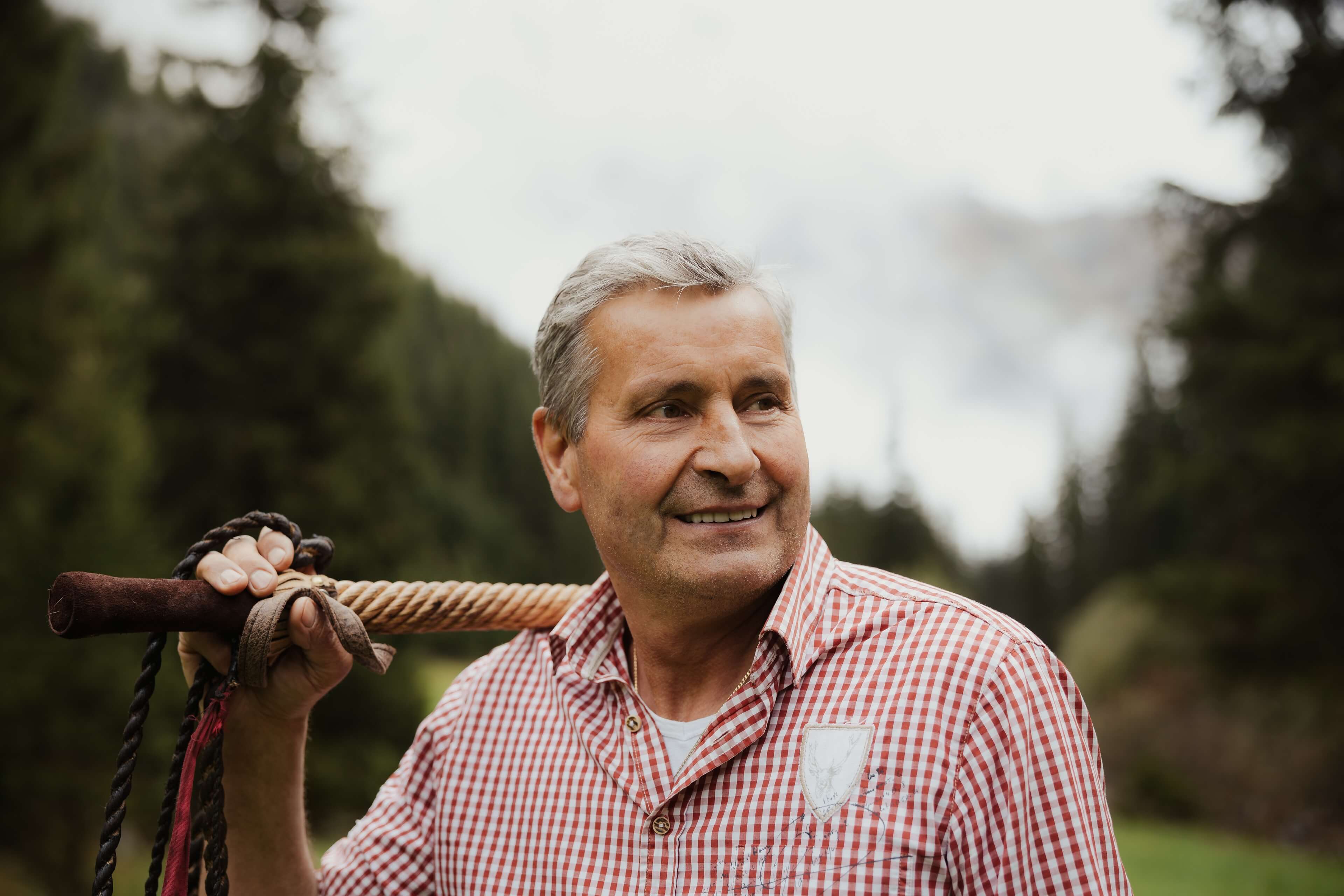 Adalbert Braunhofer con attrezzo da lavoro sulla spalla e bosco sullo sfondo