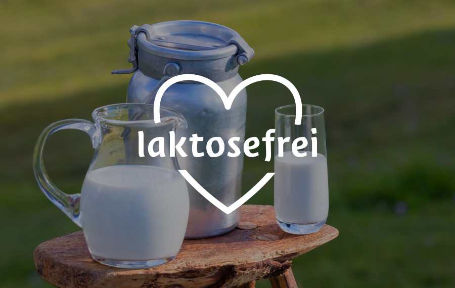 Krug und Glas Milch auf einem Holztisch im Freien, mit einem Metalleimer und der Aufschrift "laktosefrei" und einem weißen Herz darüber.