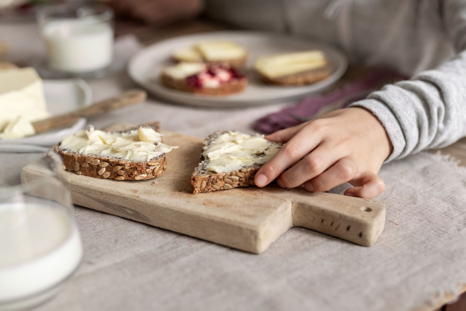 Eine Hand nimmt eine Scheibe Brot mit Streichkäse auf einem hölzernen Schneidebrett.