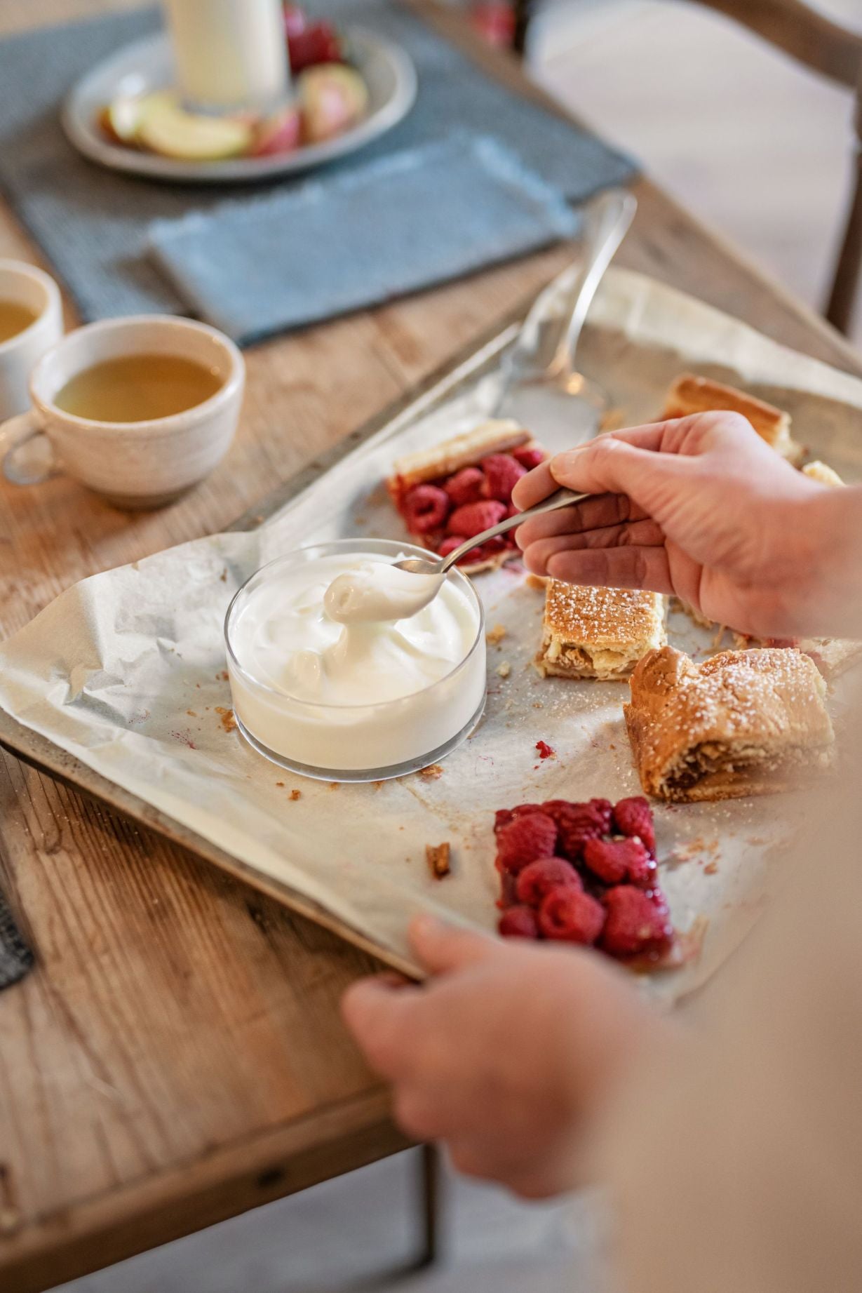 Eine Person nimmt mit einem Löffel Joghurt aus einer Glasschale, während sie ein Tablett mit Kuchenscheiben und Himbeeren in der Hand hält.