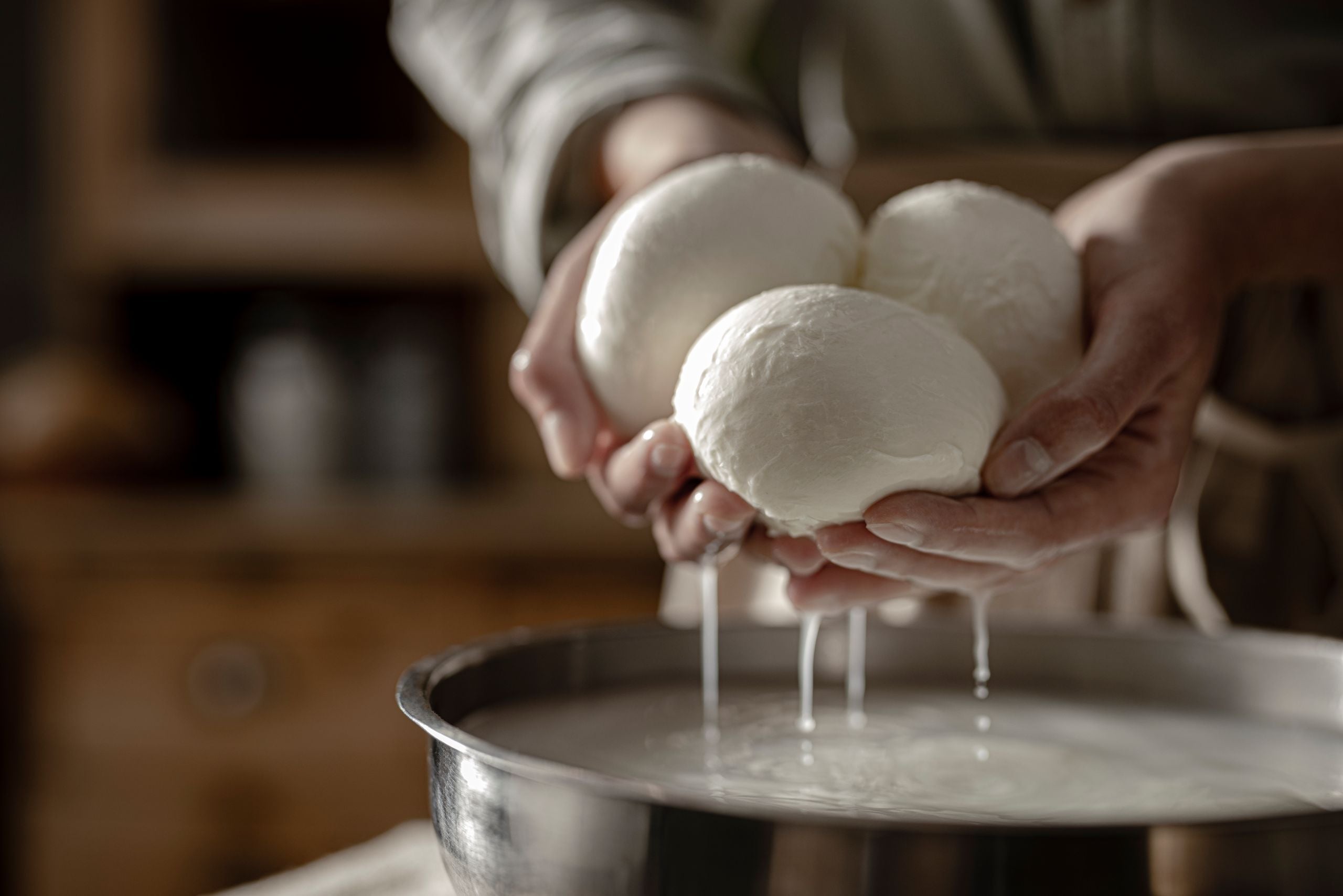 Hände heben frische Mozzarellakugeln über eine Wasserschüssel bei der handwerklichen Herstellung.