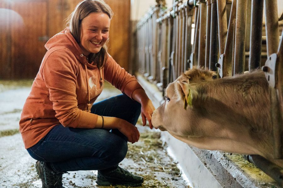 Eine Frau hockt in einem Stall und lächelt, während sie ein Kalb streichelt.