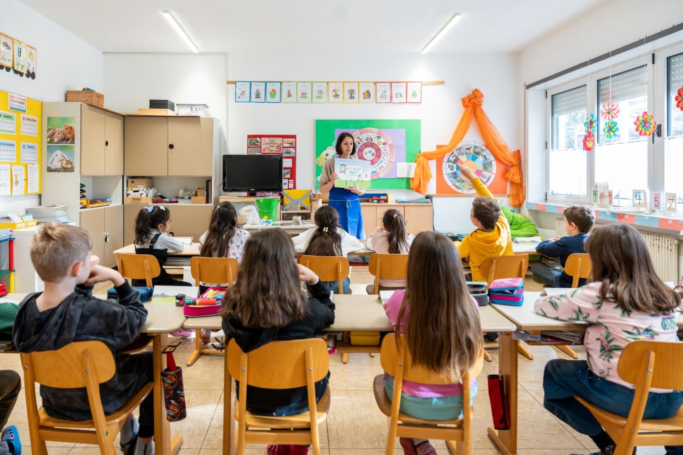 Ein stehender Lehrer zeigt einer Gruppe von Kindern, die in einem Klassenzimmer an ihren Tischen sitzen, ein Bilderbuch.