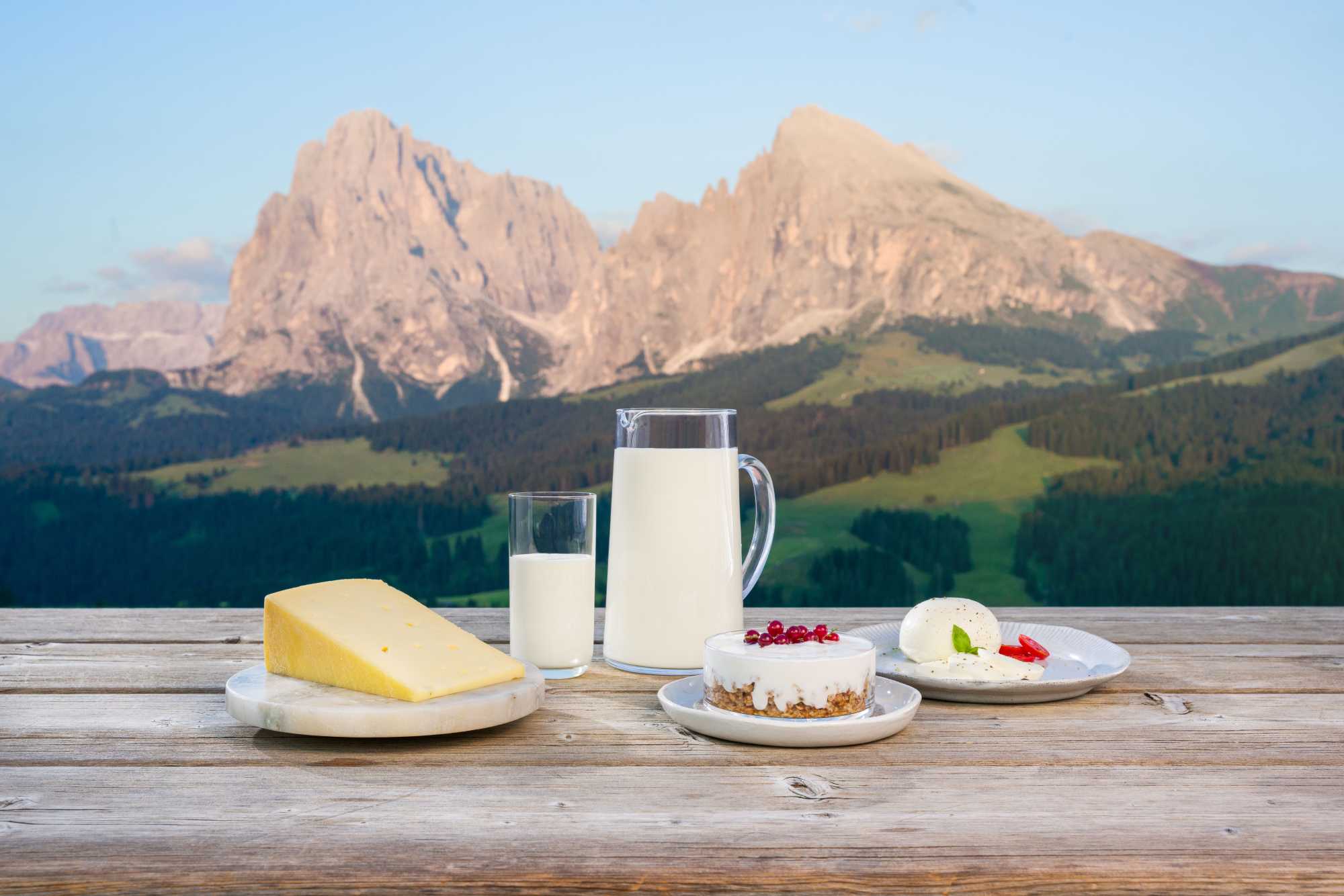 Verschiedene Käsespezialitäten wie Käse, Milch und Süßigkeiten werden auf einem Holztisch im Freien mit den Bergen im Hintergrund serviert.