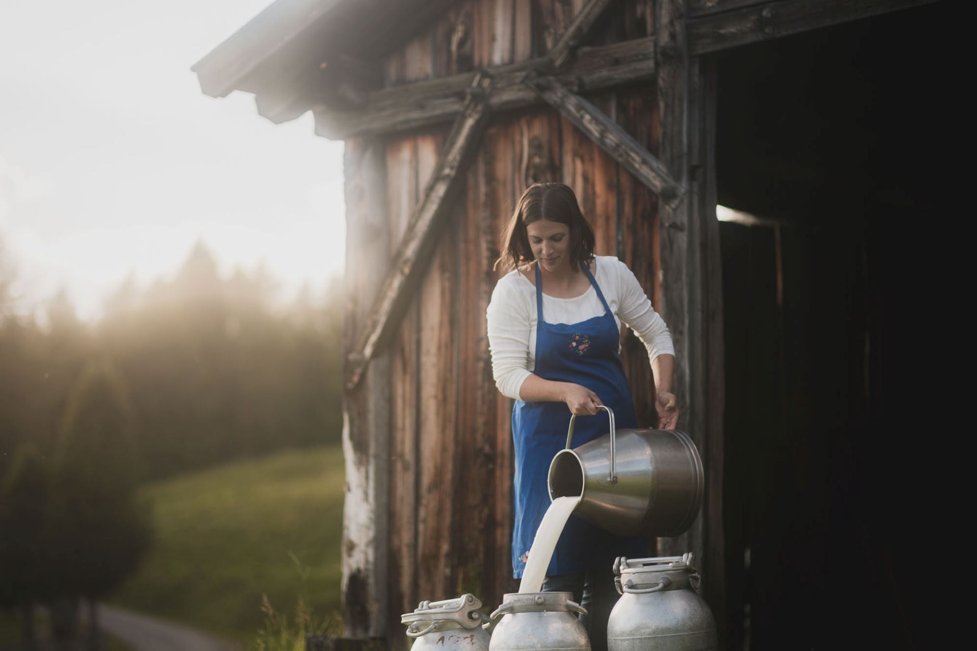 Eine Frau in einer Schürze gießt draußen vor einer Holzscheune Milch aus einer Kanne in einen Metalleimer.