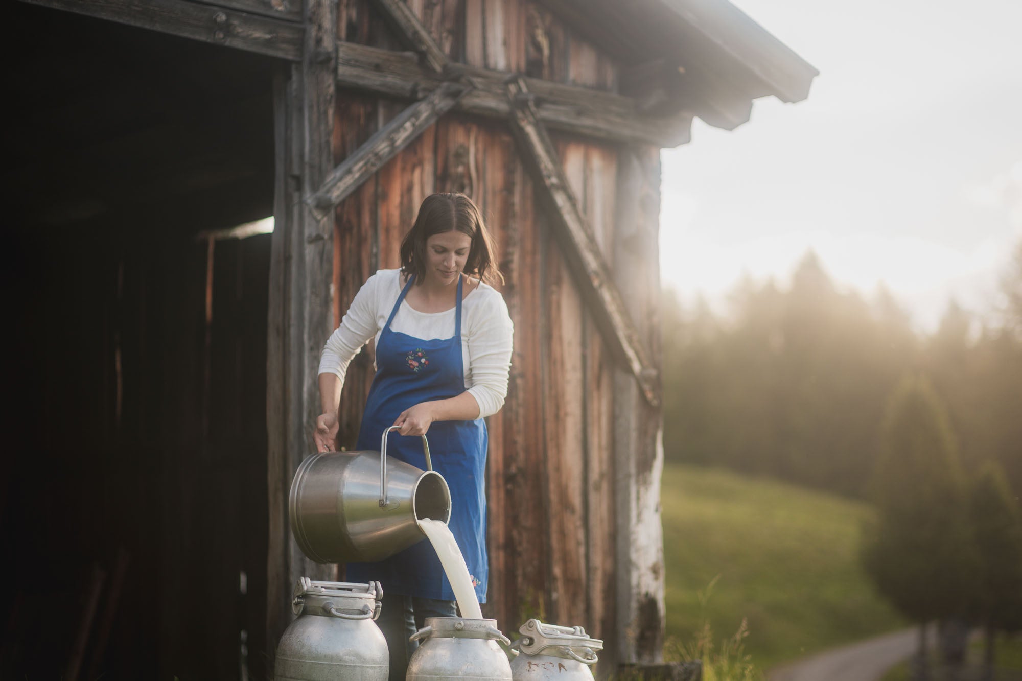 Frau in blauer Schürze gießt frische Milch aus einem Eimer in eine Milchkanne vor einer Holzscheune in den Bergen.