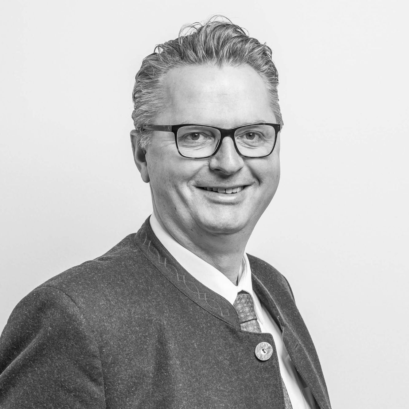 Schwarz-Weiß-Porträt von Joachim, einem Mann, der einen traditionellen Anzug mit einer verzierten Jacke und einer Brille trägt, während er lächelt.