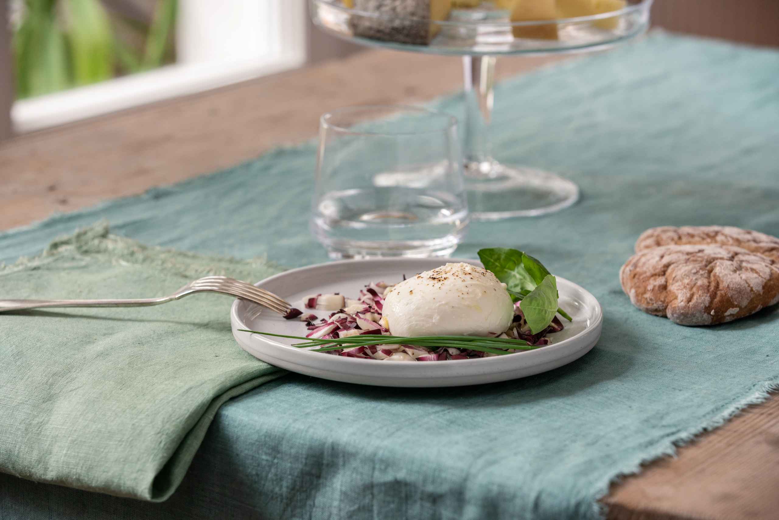 Ein weißer Teller mit frischem Mozzarella und einem aromatischen Kraut, im Hintergrund ein Laib Brot und ein Glas Wasser.