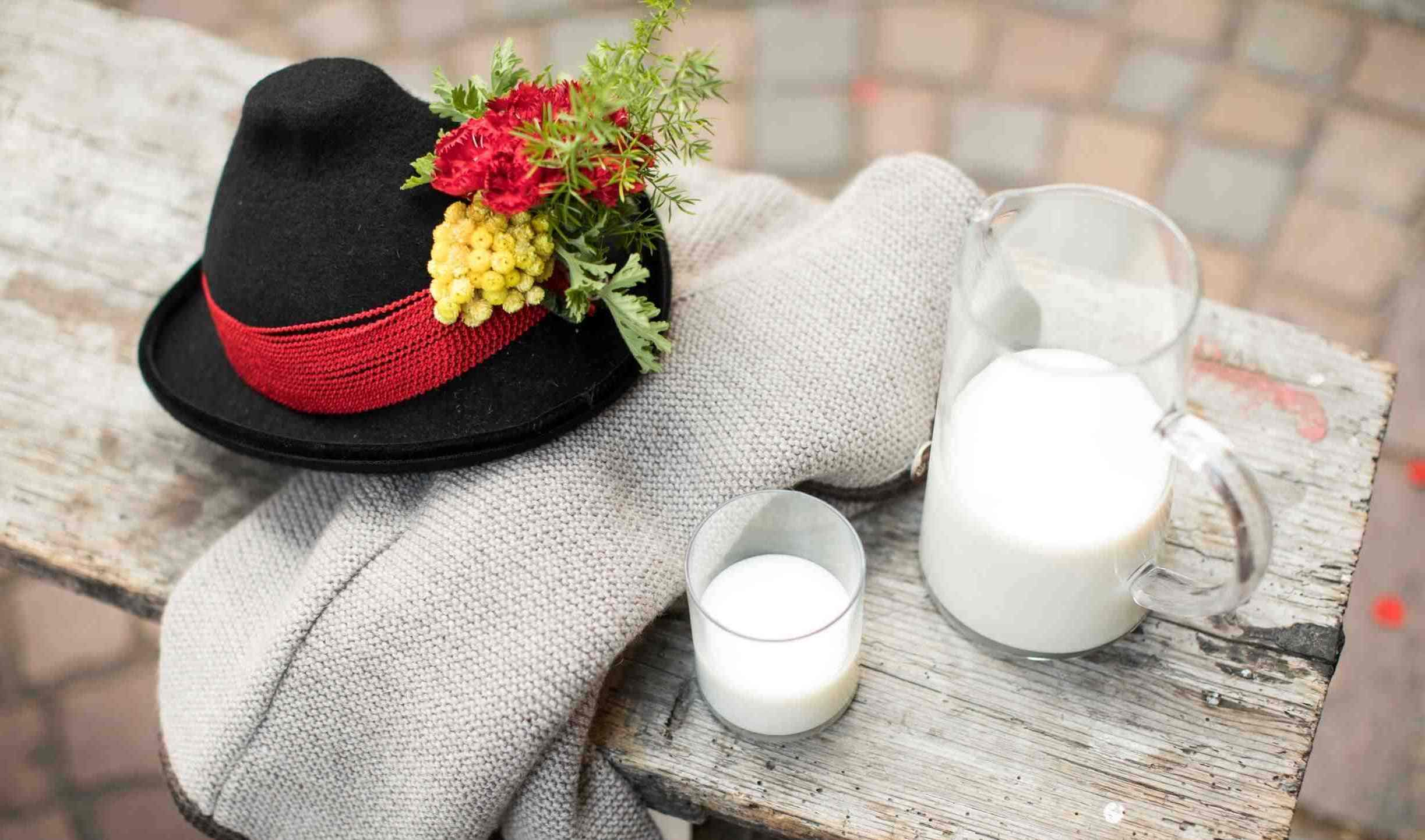 Glas und Kanne Milch neben einem traditionellen Hut mit Blumen, auf einer Holzbank.