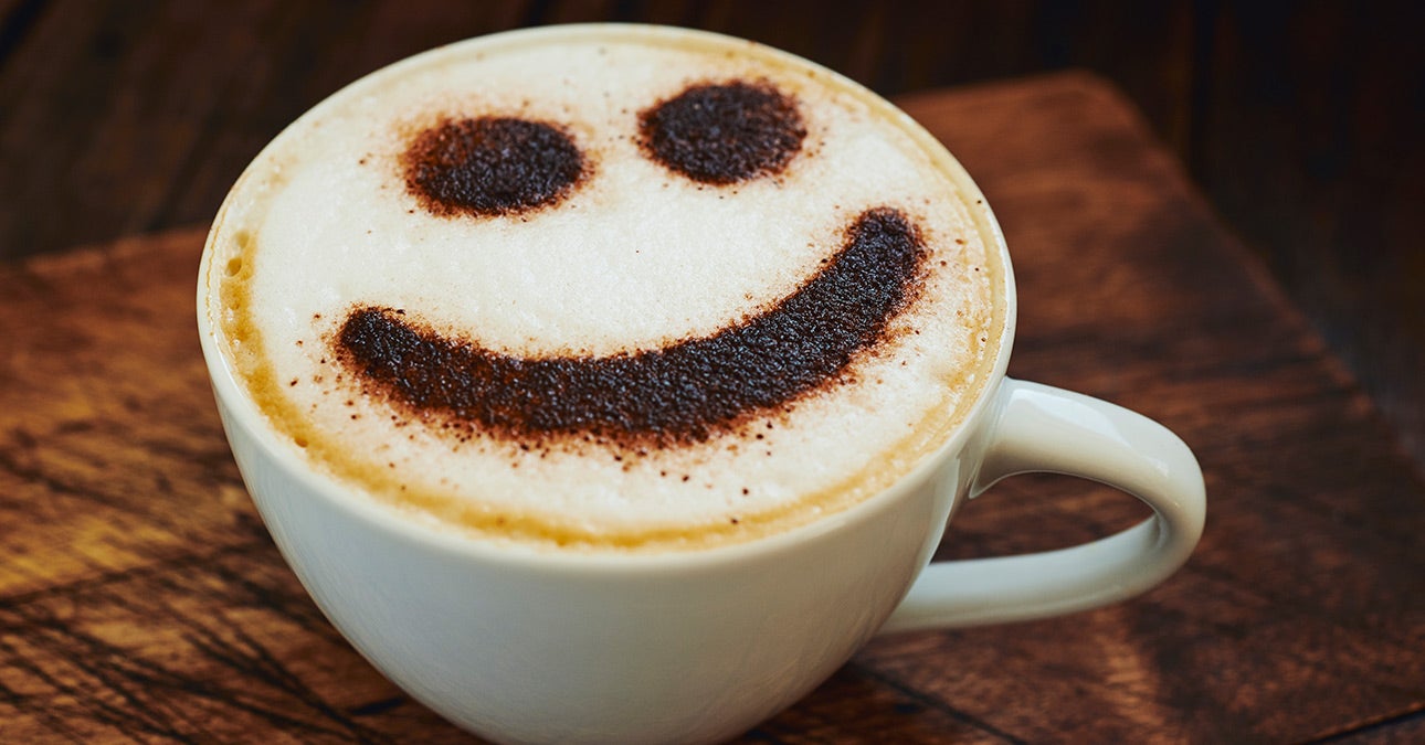 A cappuccino in a white mug on a wooden table. It is dusted with chocolate powder in the shape of a smiley face, celebrating International Week of Happiness at Work. 