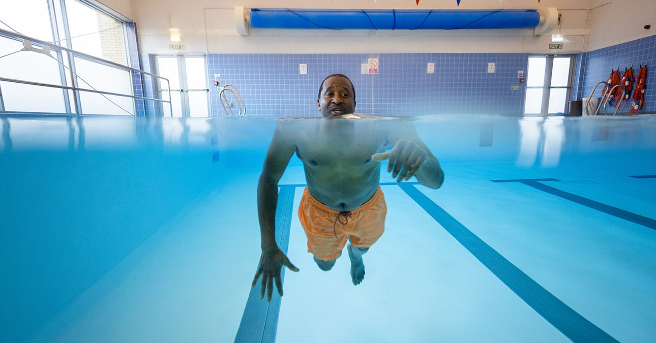 The photo is split horizontally, the bottom half underwater in a swimming pool and the top half above the water. A man in orange swimming shorts is keeping fit by swimming towards the camera, his head out of the water and the rest of his body beneath the waterline.