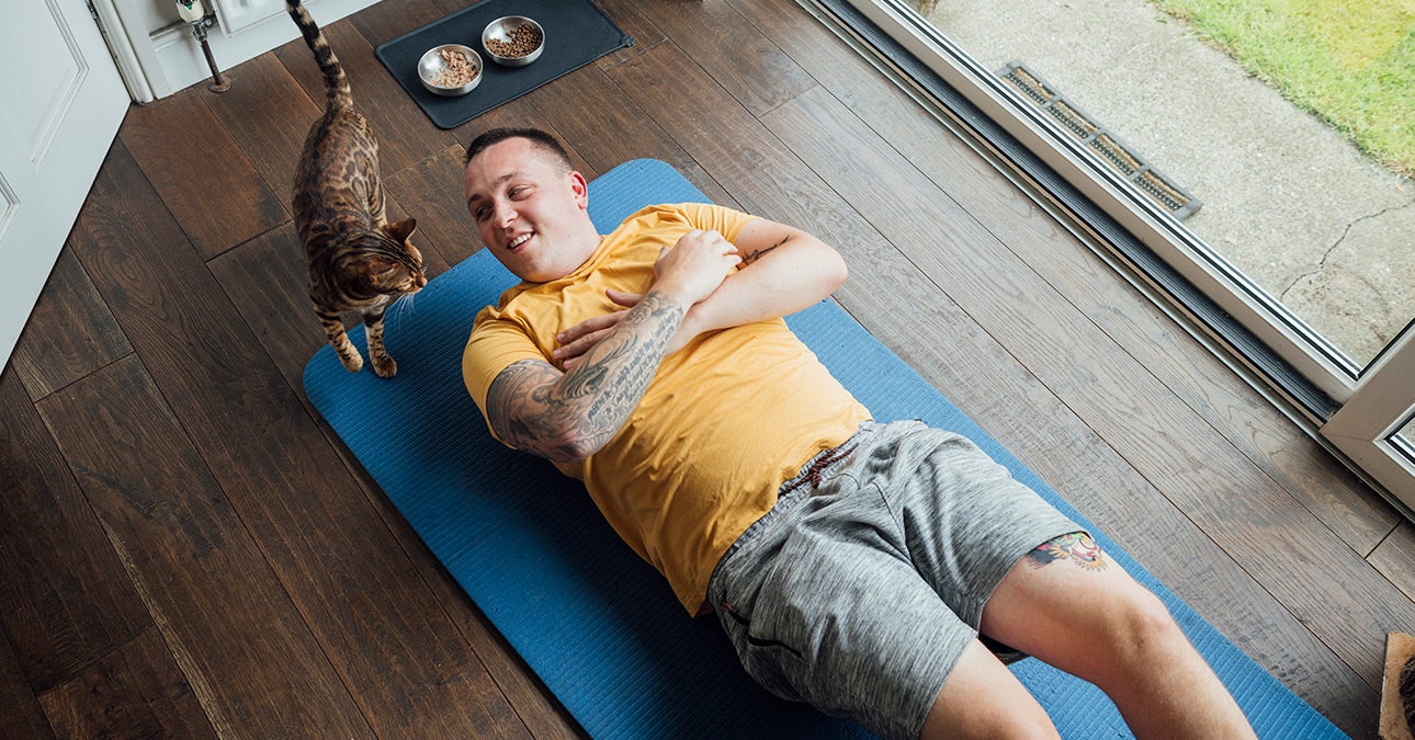 A man in grey shorts and a yellow t-shirt lies on his back on a blue yoga mat on the floor. His arms are folded across his chest. He appears to have been interrupted from meditating, practising mindfulness or doing yoga by his tabby cat, which has appeared near his head. He smiles at the cat.