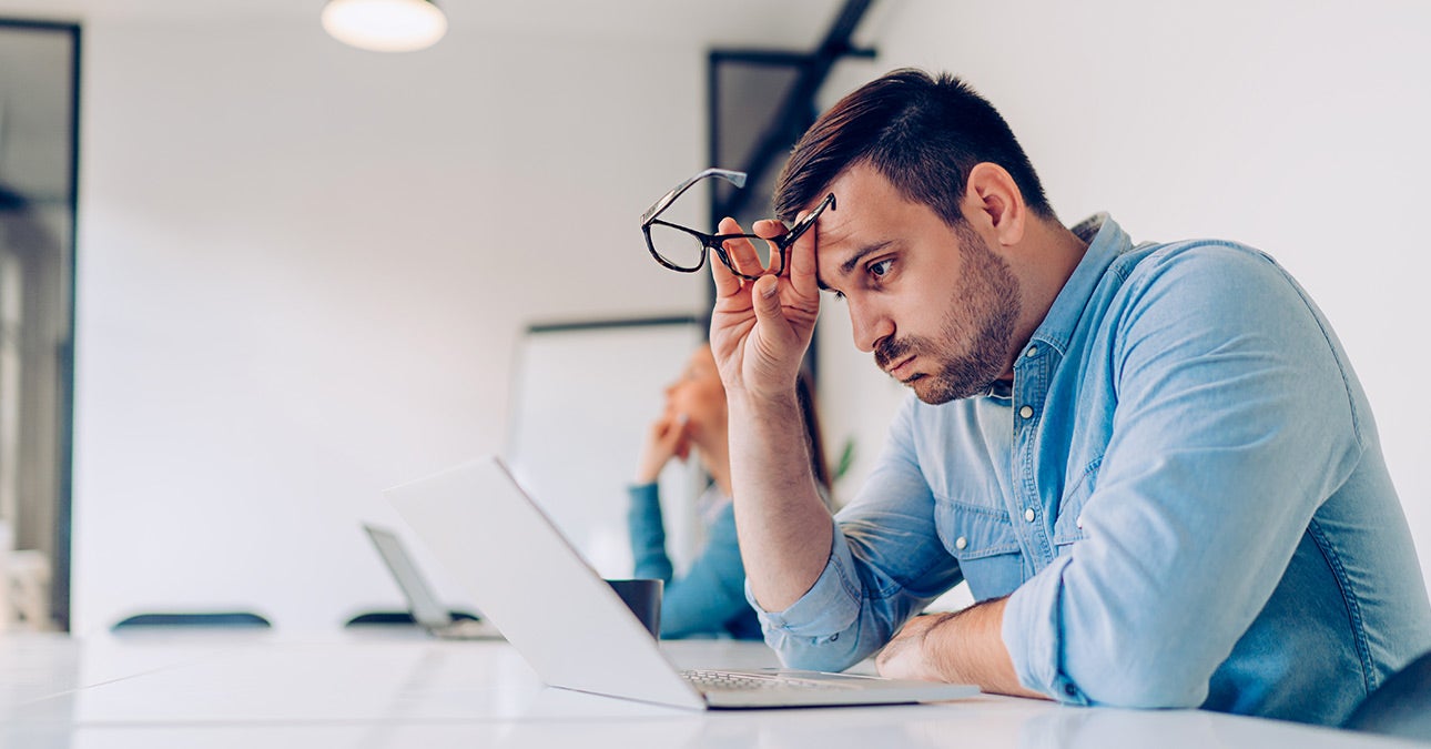 Stressed man in blue shirt in front of a laptop. He has removed his glasses and is propping his head on his hand as he stares at his computer.