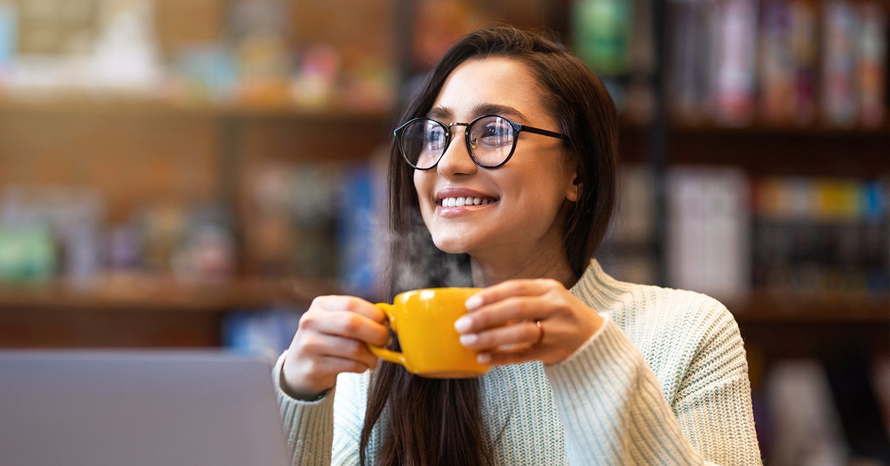 Woman at her laptop wearing glasses smiles while holding a yellow cup of a hot drink giving off steam.