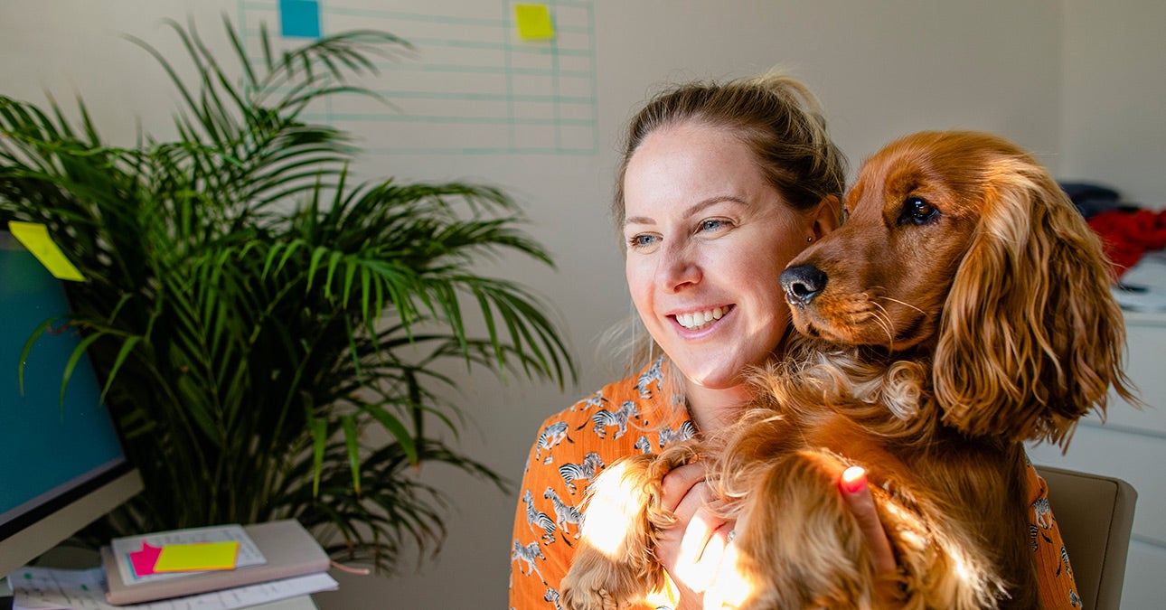 Smiling woman working from home with her spaniel in her lap. She is a hybrid worker and using the new freedoms of this lifestyle to balance her home and work worlds.