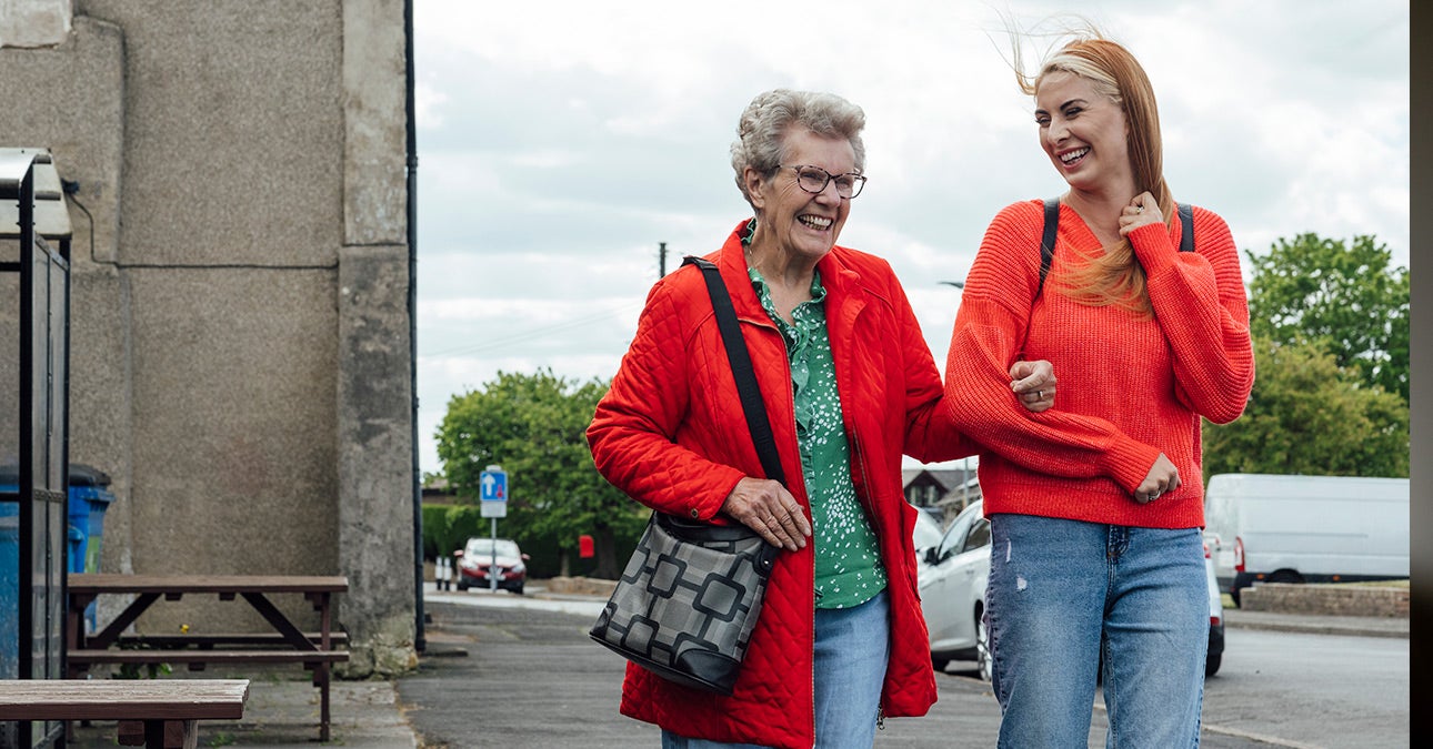 Two women dressed in jeans and red tops. The woman on the left is an older woman with glasses. She is steadying herself by taking the arm of the younger woman on the right.
