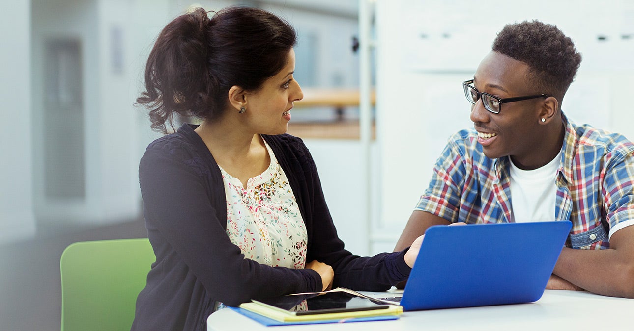 A woman and a man sit at a round table in an office setting. The woman has a notepad and a blue laptop open on the desk. She wears a black cardigan and a white top with small flowers on it. The man wears a blue, red and yellow checked shirt unbuttoned over a white t-shirt. He has glasses. They are discussing what is on the laptop screen: a plan to support employees with neurodiversities.