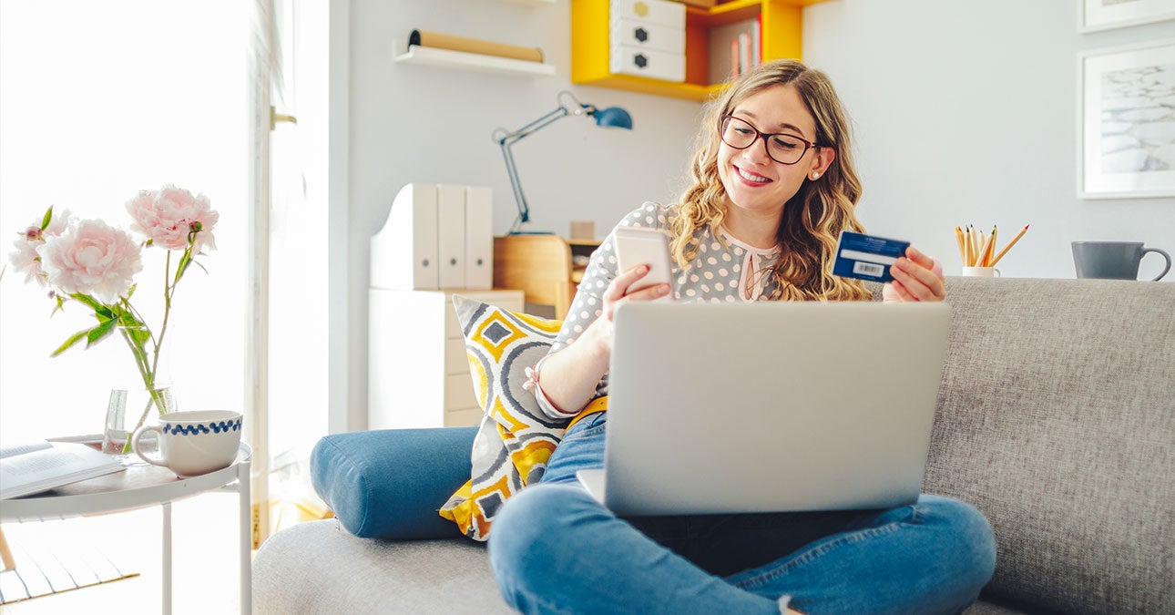 A woman sits on a couch and holds a credit card and a phone