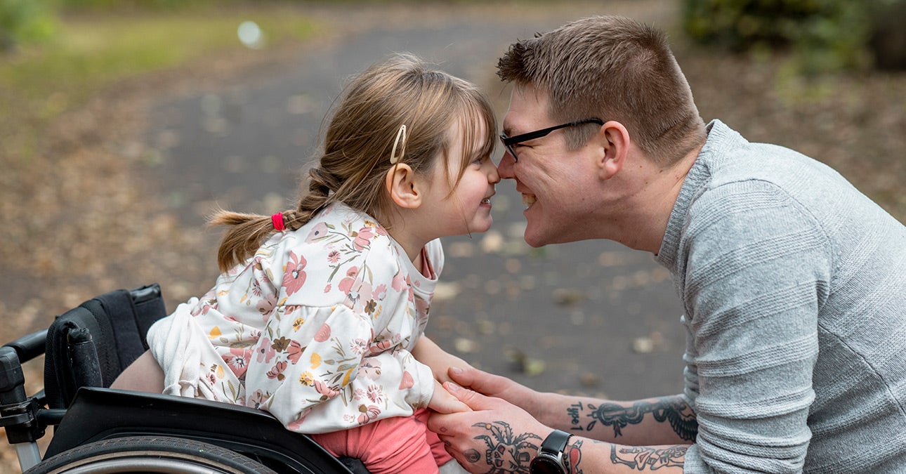 As a carer, a father in a grey jumper and glasses crouches down to touch noses with his young daughter. His daughter is a wheelchair user dressed in pink trousers and a white shirt with floral pattern. Image from Unum to highlight Carers Week 2023.