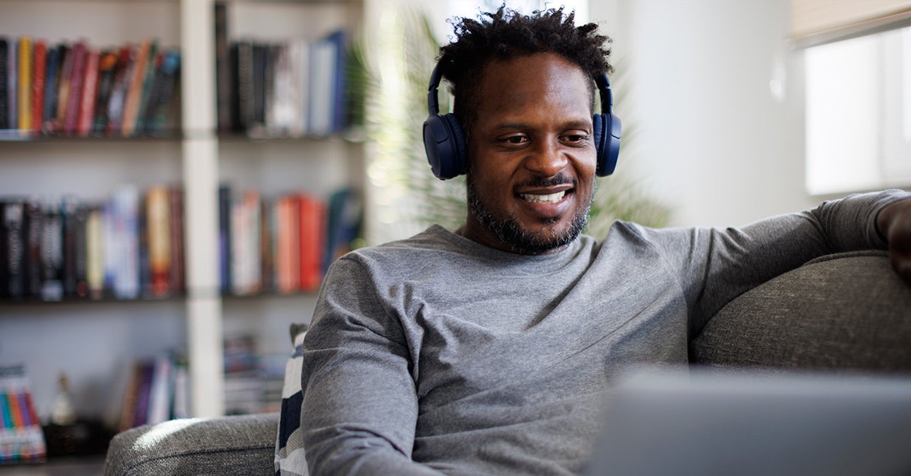 A man sits in his living room with his headphones on and laptop open, browsing podcasts and videos as part of Unum's wellbeing resources. He smiles.