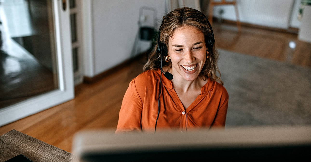 Woman working from home in orange blouse using her headset to participate in a work video call. Her employer offers Wellbeing Checks from Unum to ensure she remains connected and well while working remotely.