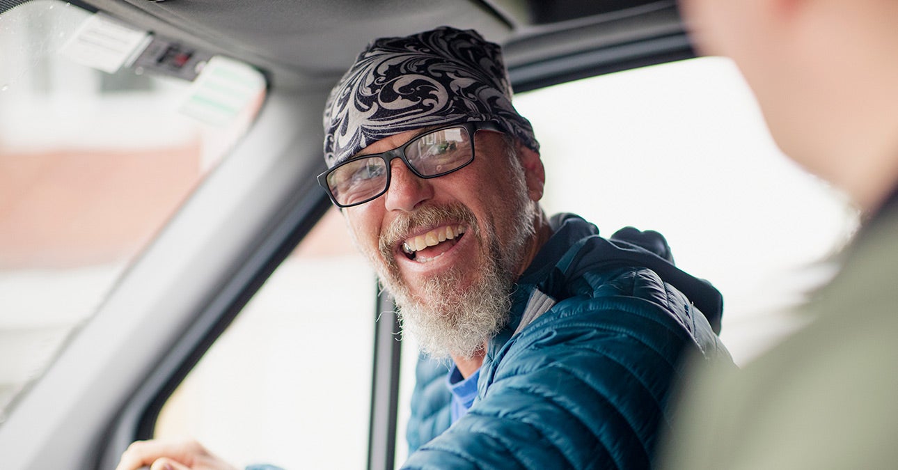 A smiling man with a beard and glasses is at the wheel of a van and turning to his colleague in the passenger seat. The conversation surrounds men's mental health, a topic that's often taboo for men but one Unum wishes to highlight as crucial for Men's Health Week.