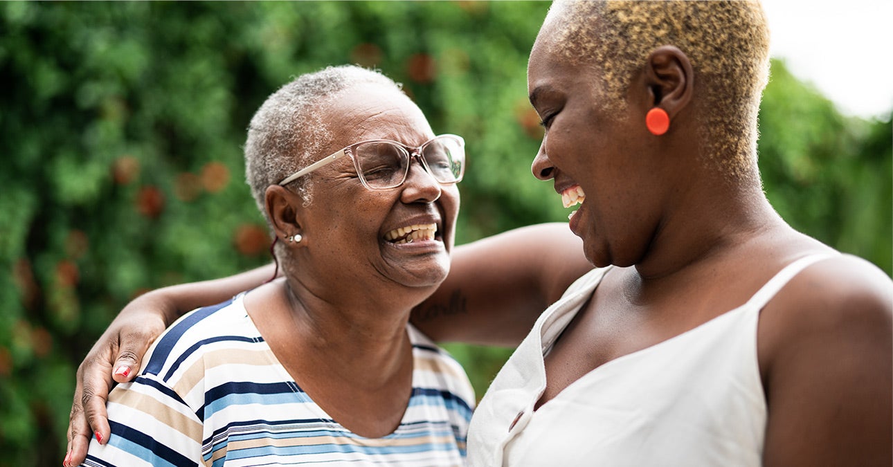 A smiling daughter and mother. The mother is an older person in glasses and with grey hair. She's wearing a striped t-shirt and embracing her daughter, who is wearing a white sleeveless top. The daughter is a carer for her mother, an elder needing support, and features for Unum's article about caring for carers.