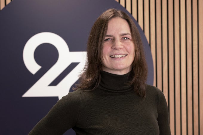 Headshot of Liz Reynolds, with the 26 logo in the background.