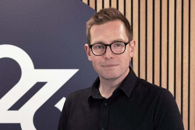 Headshot of Alex Blaikley, with the 26 logo in the background.