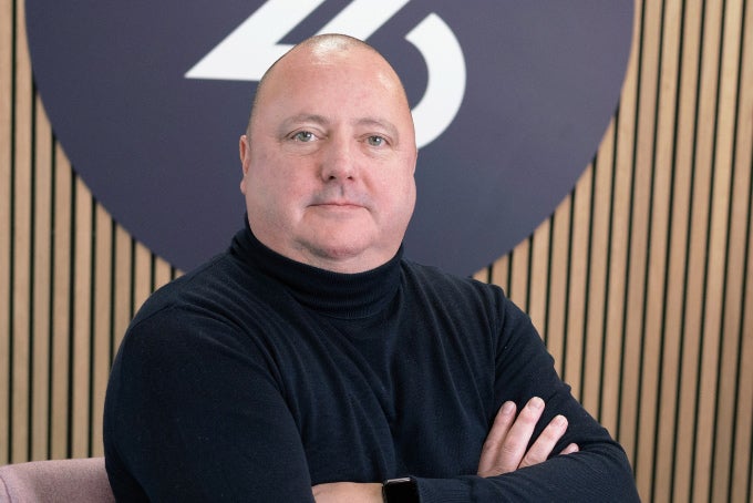 Headshot of Adam Powers, sat on a chair, arms crossed, with the 26 logo in the background.