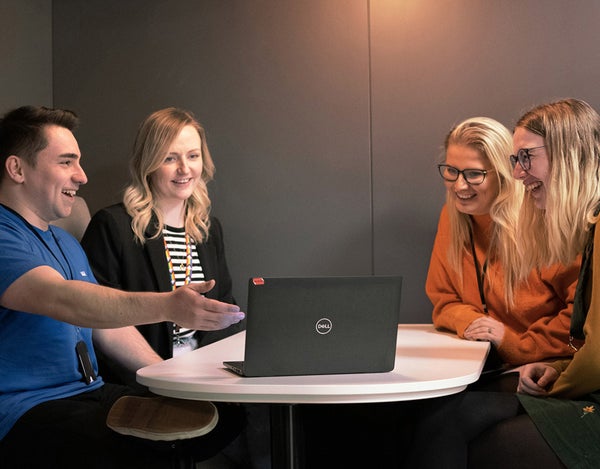 Four 26 employees sat around a table, talking, smiling and laughing. They are all looking at a laptop, on the table, and one of the employees is pointing at it.