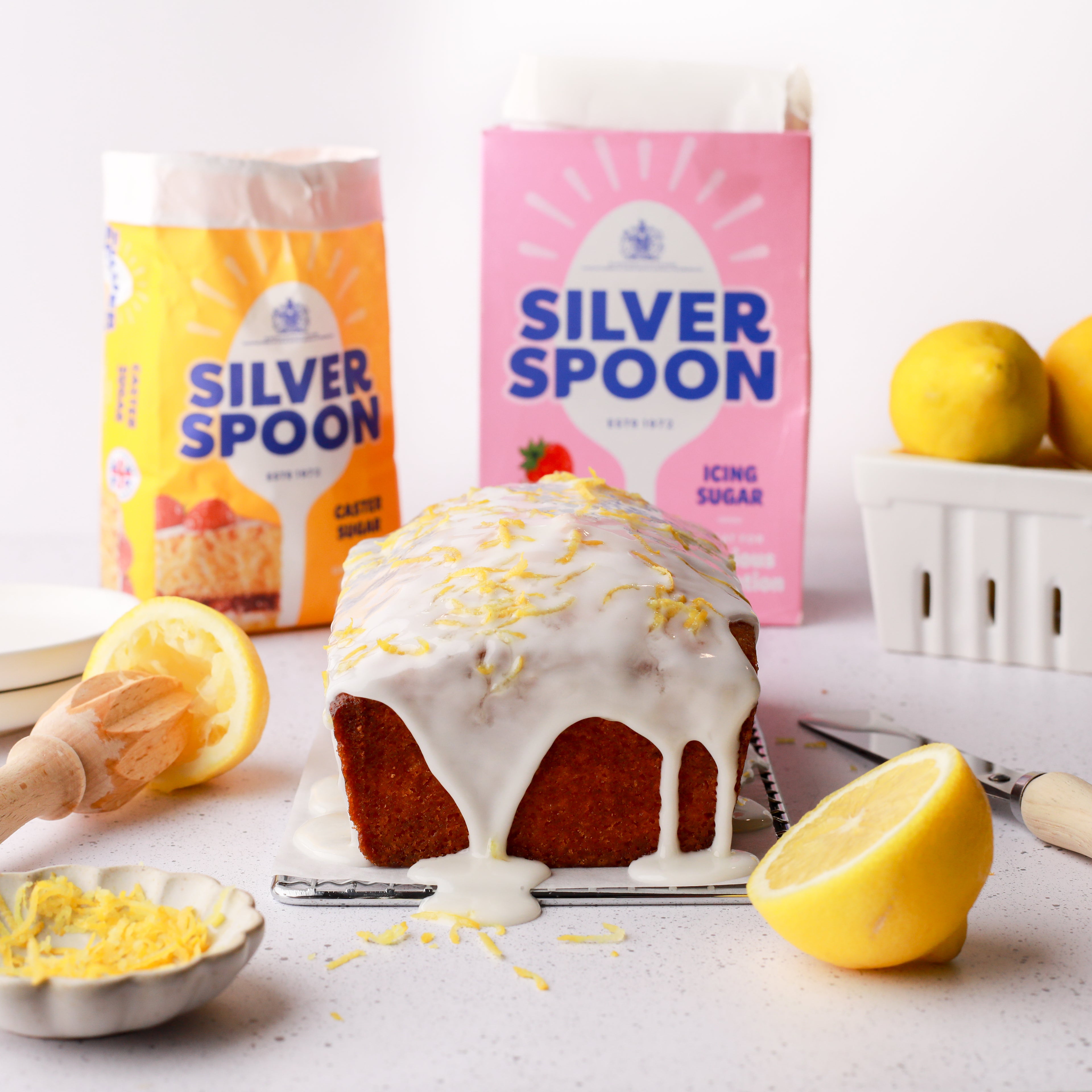 A freshly made lemon drizzle cake surrounded by lemons in front of the yellow and pink packs of Silver Spoon caster and icing sugar.