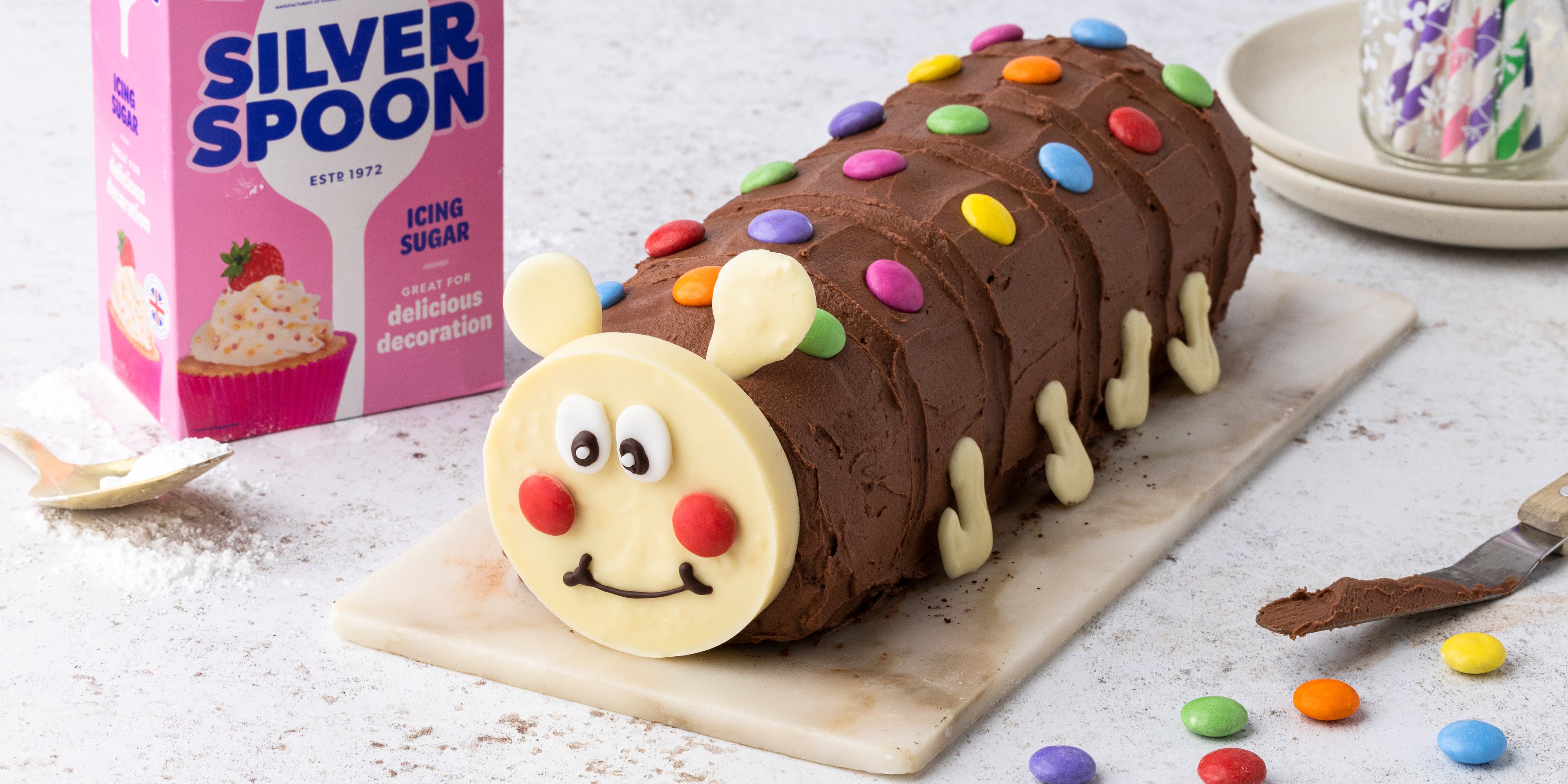 Caterpillar cake decorated with smarties on a marble tray next to a pink pack of Silver Spoon icing sugar.