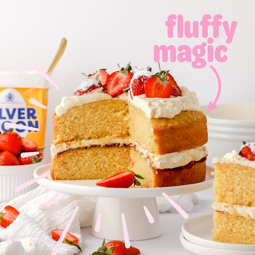 A freshly made Victoria Sponge cake on a cake plinth, topped with strawberries and buttercream with a caption saying "fluffy magic" and pointing to the cake.