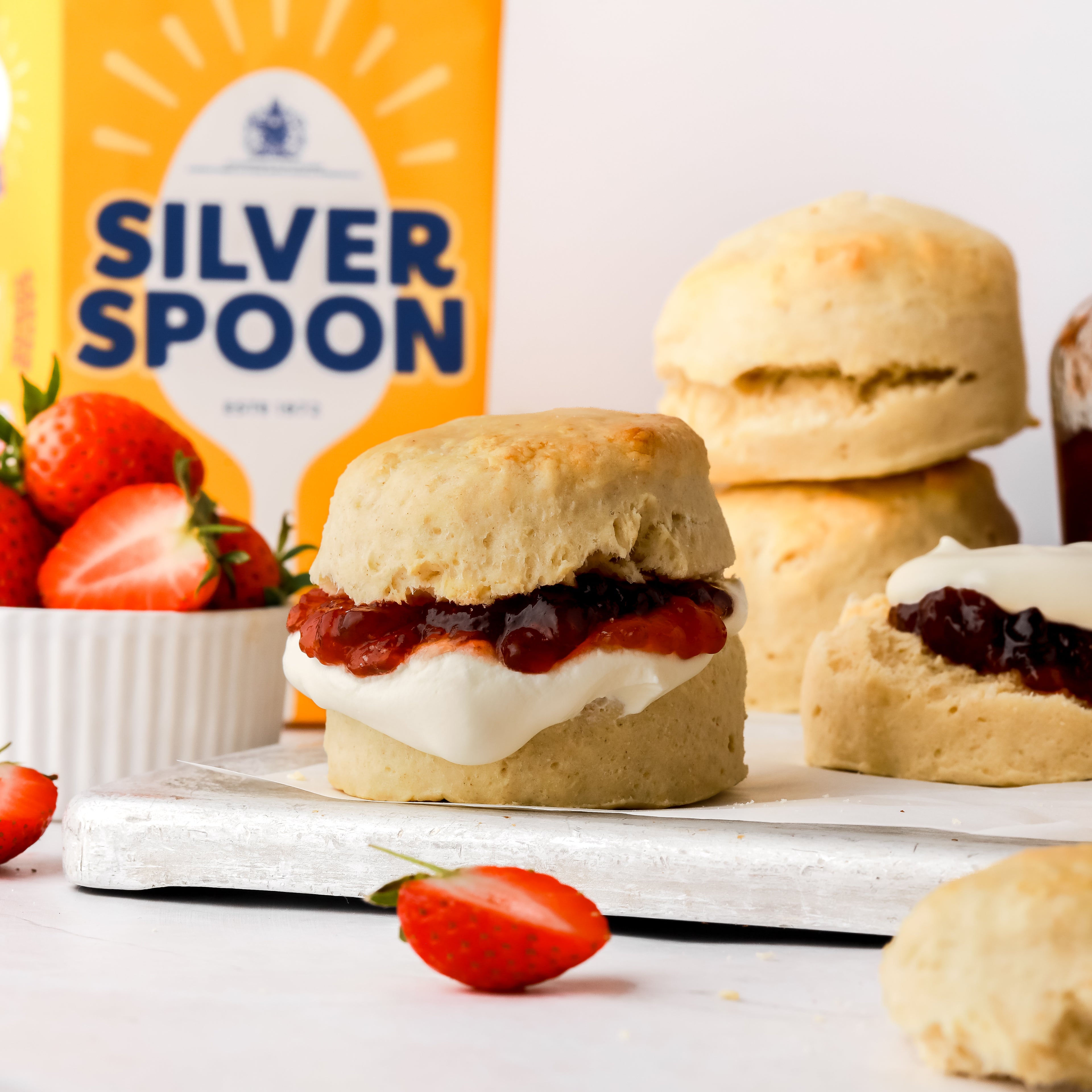 Freshly made scones with a small pot of sliced strawberries in front of a yellow bag of Silver Spoon caster sugar.
