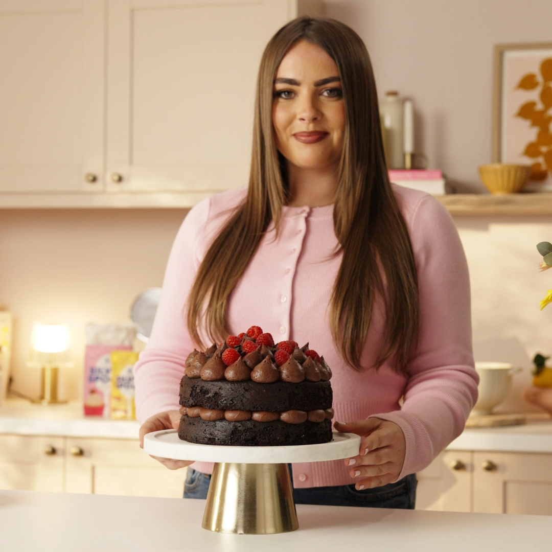 Poppy Carr standing with her freshly made chocolate cake that's decorated with chocolate buttercream and raspberries.