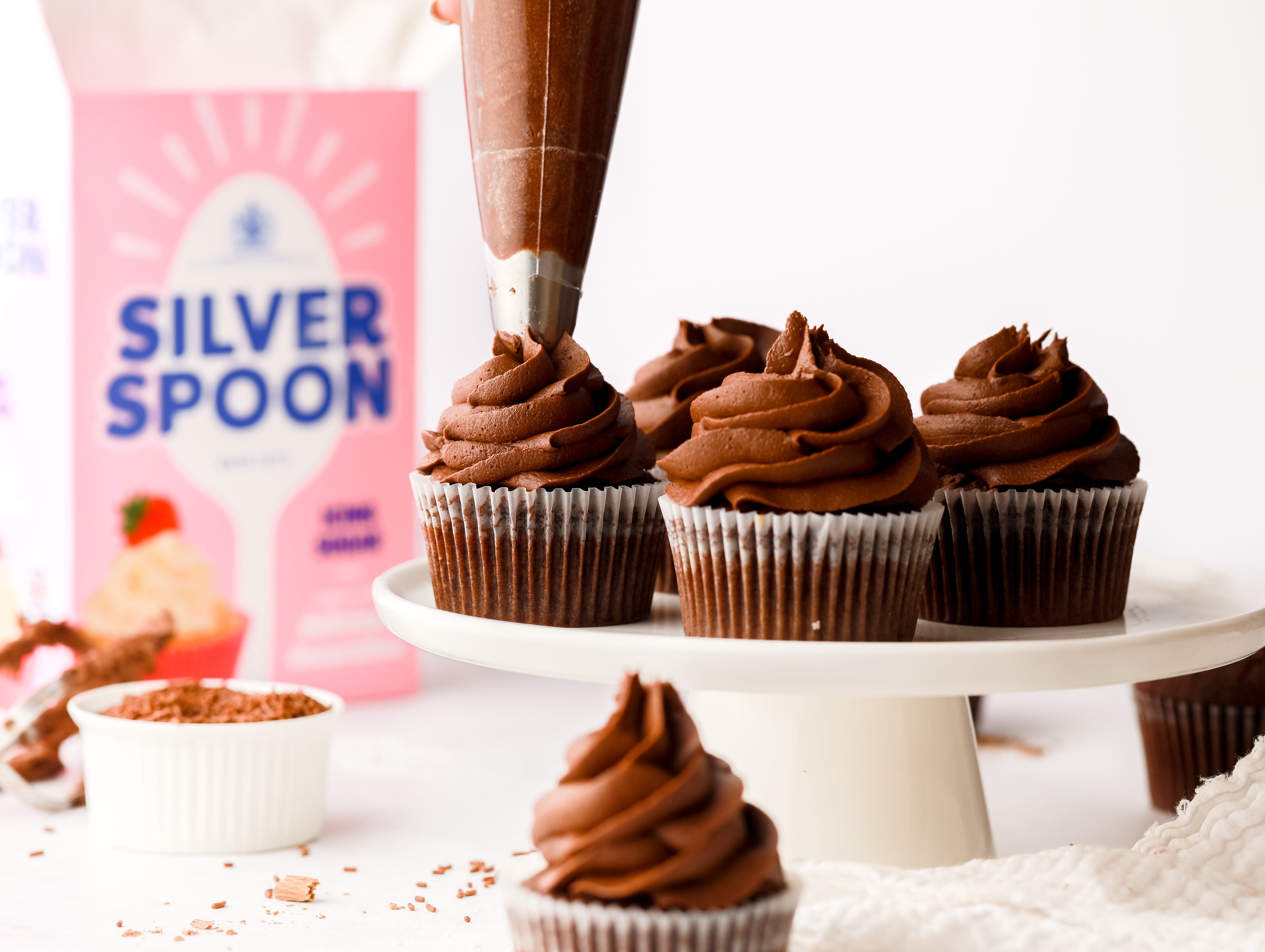 A cake plinth of chocolate cupcakes with swirled chocolate icing on top in front of a pink pack of Silver Spoon icing sugar.