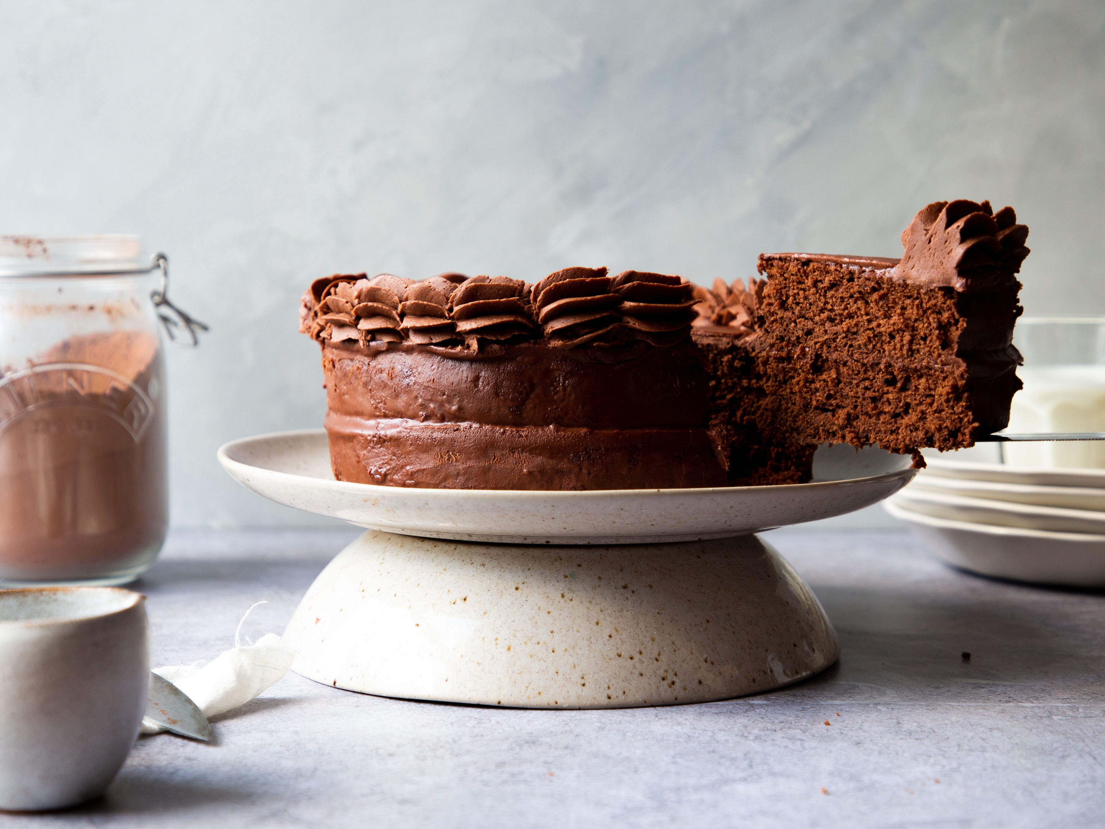 Classic Chocolate Cake on a cake stand with a slice being taken out of it held by a knife. Next to a jar of cocoa powder