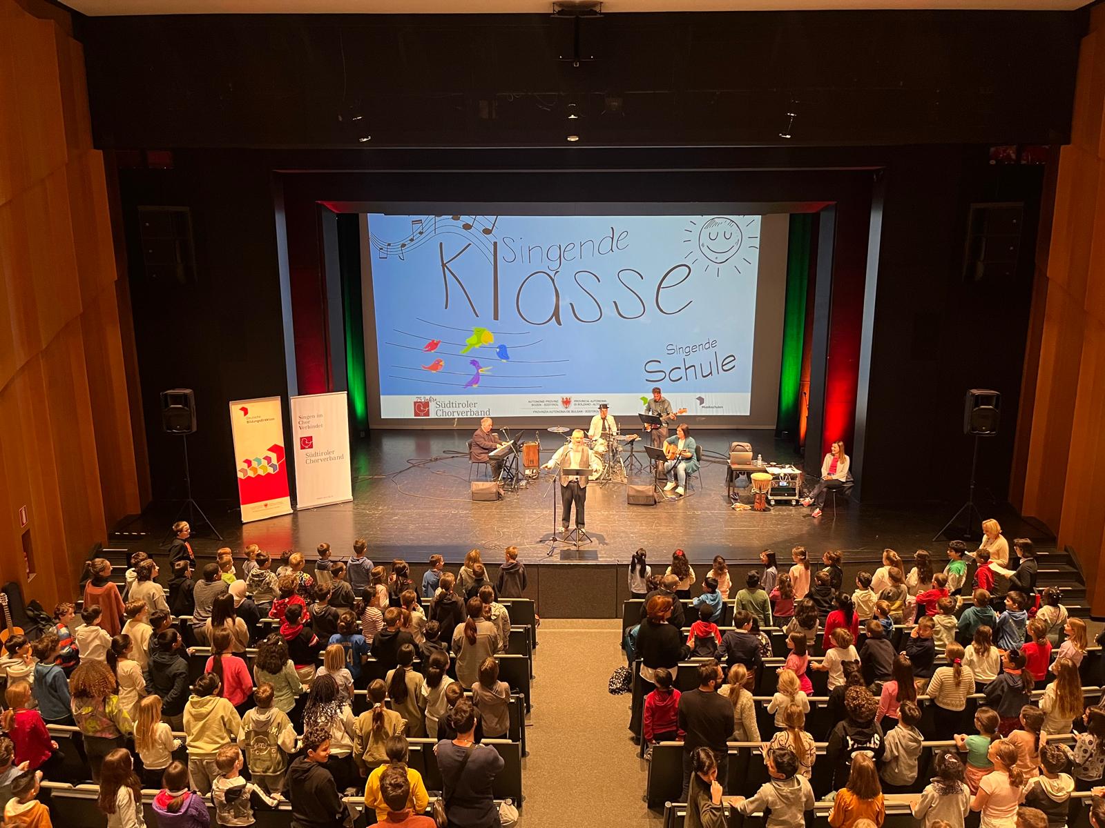 Bei den Singfesten werden die Schülerinnen und Schüler von einer besonderen Band begleitet; dazu gehören Andreas Benedikter (Klavier und Flöte), Manfred Gampenrieder (Perkussion) Barbara Prantl (Gitarre) und Rottensteiner Armin (Bass); in der Bildmitte: Hanno Deluggi, rechts im Bild: Sigrun Falkensteiner