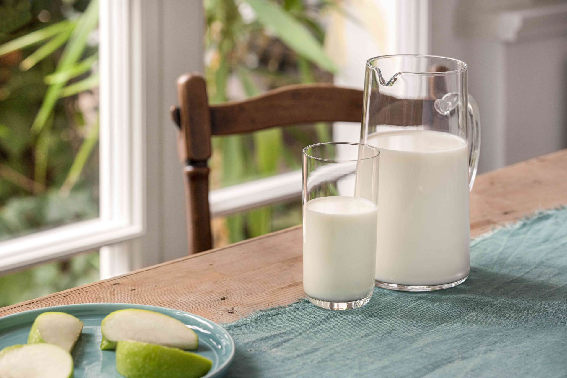 Karaffe und Glas mit frischer Milch auf einem Tisch mit blauem Tuch neben einem Teller mit grünen Apfelscheiben.