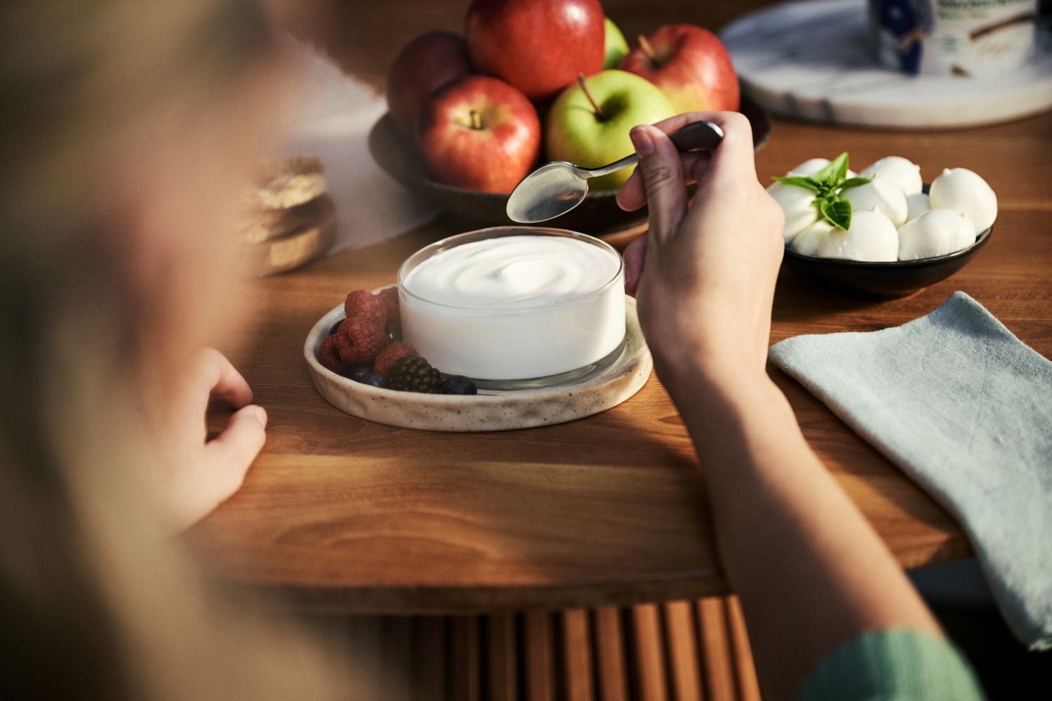 Eine Hand taucht einen Löffel in eine Schale mit Joghurt und Beeren, auf einem Holztisch stehen eine Schale mit Mozzarella und eine mit Äpfeln.