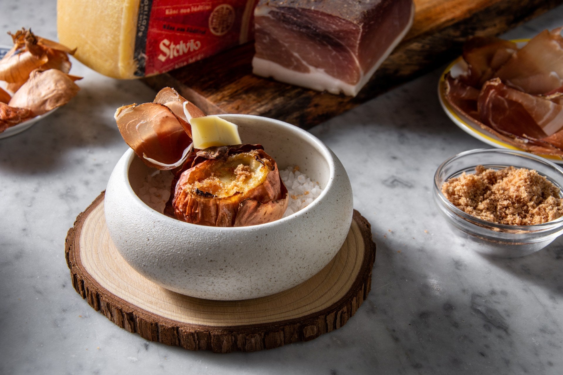Geschmorte Zwiebel gefüllt mit Stilfser Käse und Südtiroler Speck g.g.A. Pulver, in einer Schale auf grobem Salz serviert, garniert mit Speck und Käse.