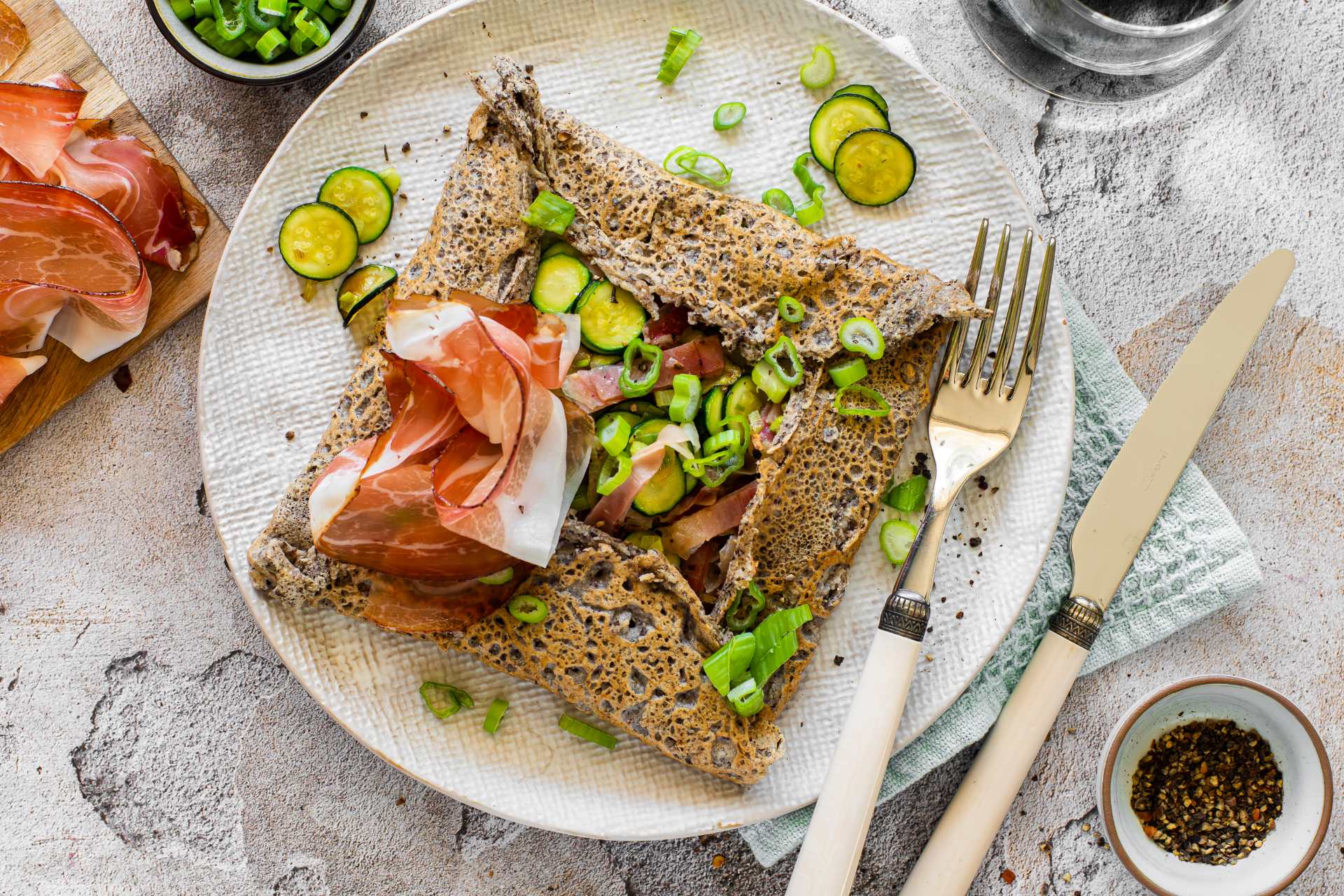 Bretonische Galette aus Buchweizen, gefüllt mit Südtiroler Speck g.g.A. und Zucchini, auf einem Teller serviert, garniert mit Zucchinischeiben und Frühlingszwiebeln.