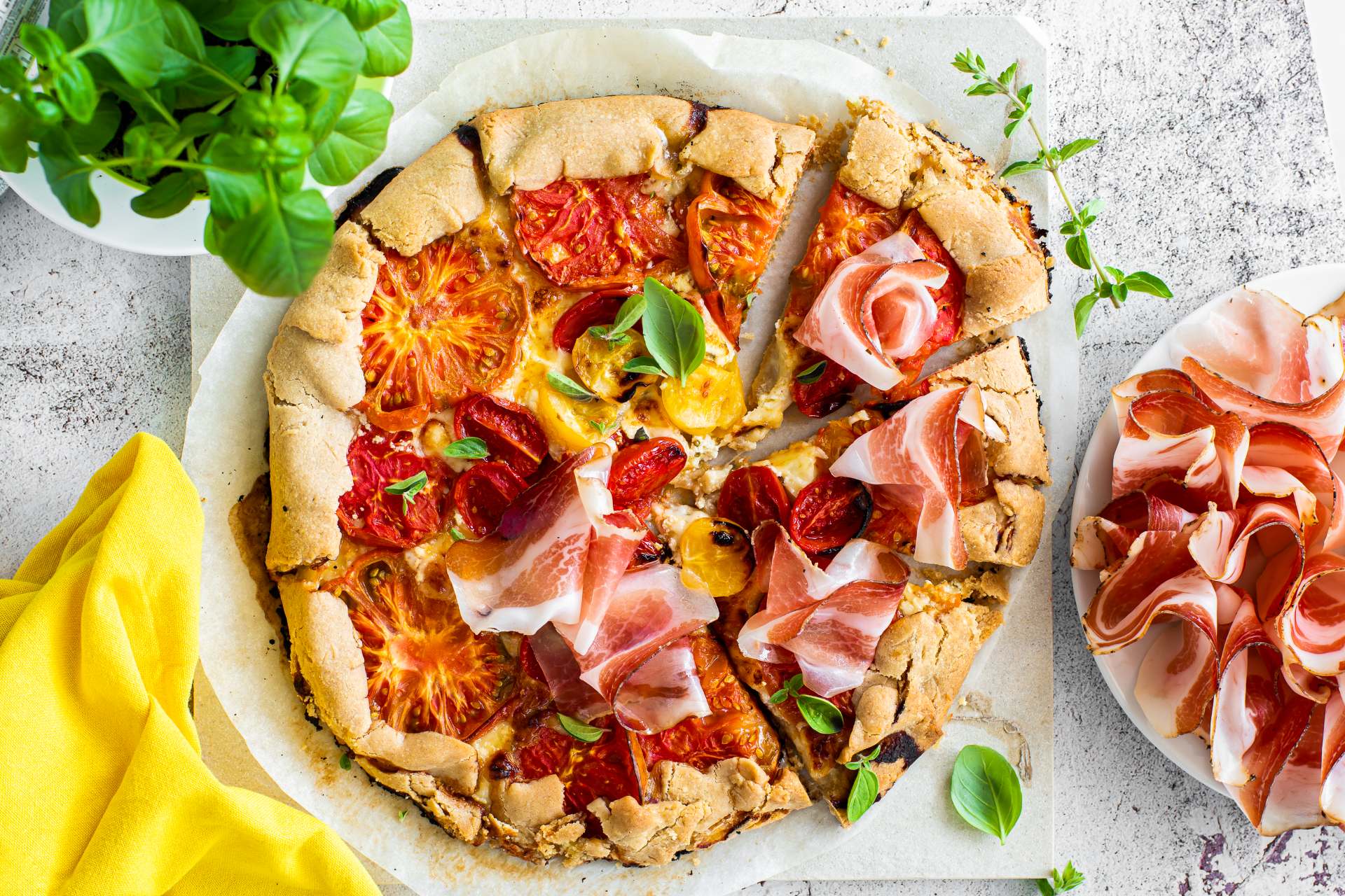 Herzhafte Galette mit rustikaler Kruste, gefüllt mit Tomaten, Südtiroler Speck g.g.A. und Basilikum, serviert mit Südtiroler Speck g.g.A. in Scheiben daneben.