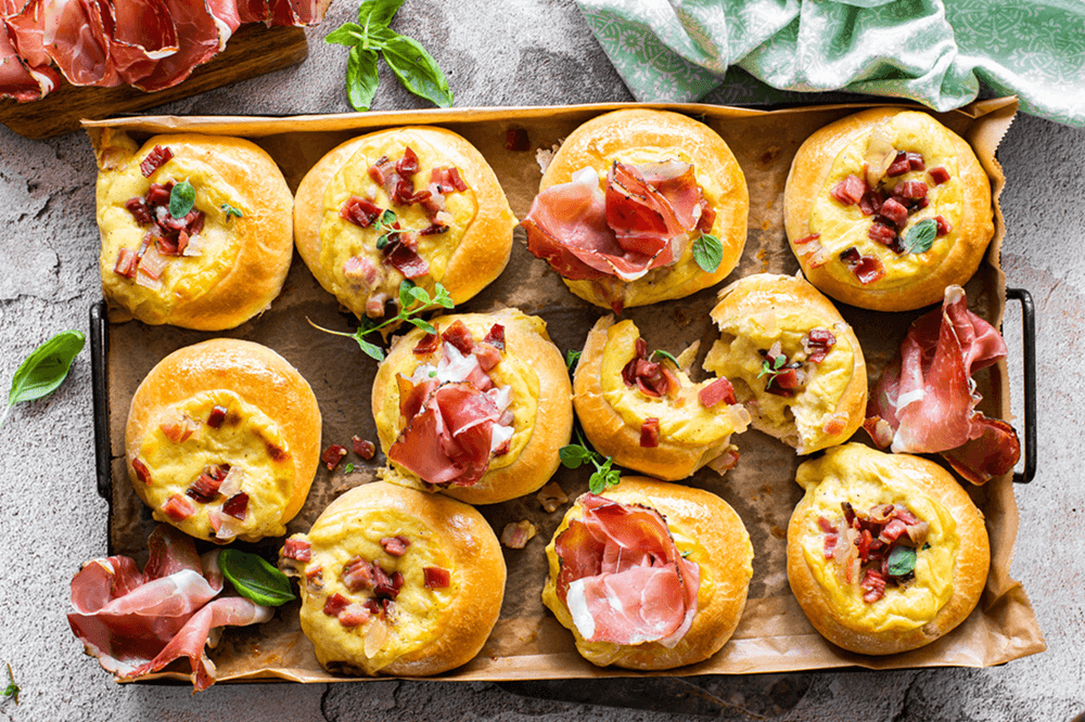 Frisch gebackene Buns auf einem Backblech, gefüllt mit Käse-Béchamelsauce und dekoriert mit Südtiroler Speck g.g.A