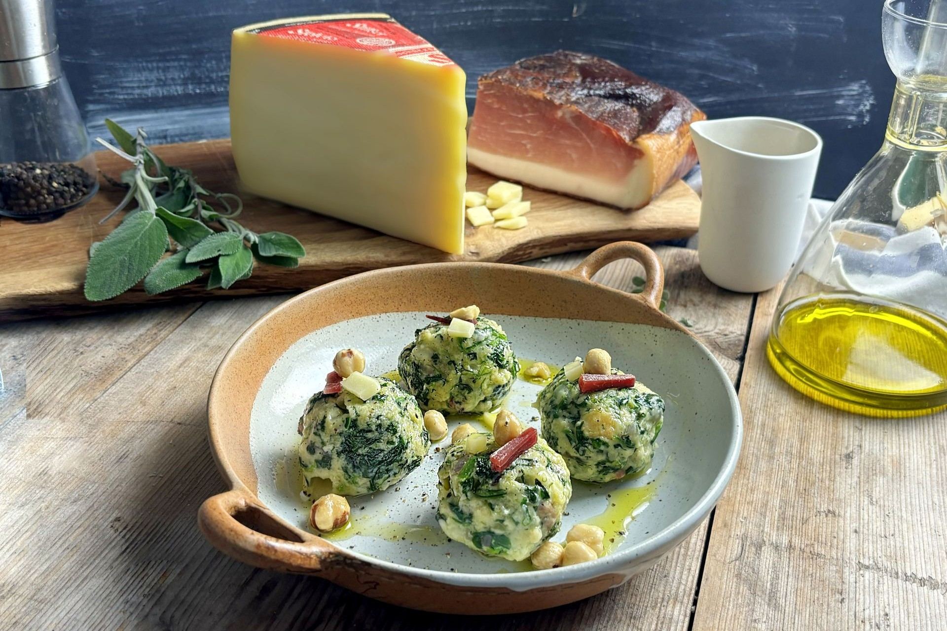 Spinatknödel mit Südtiroler Speck g.g.A. und Stilfser Käse, in einer Pfanne serviert, mit Olivenöl beträufelt, Nüssen und Käsestücken garniert.