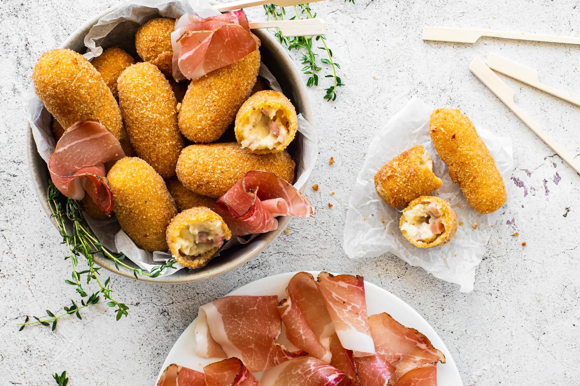 Knusprige Croquetas mit Südtiroler Speck g.g.A. in einer Schüssel, teilweise aufgeschnitten und mit cremiger Füllung, garniert mit Speckscheiben und frischen Kräutern.
